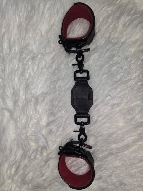Owned Riding Cuffs - Tethered 2 Black English Bridle leather Owned Riding Wrist Cuffs with red deersplit lining, connected by a matching leather tether with black swivel snap hardware, shown laid out lengthwise.