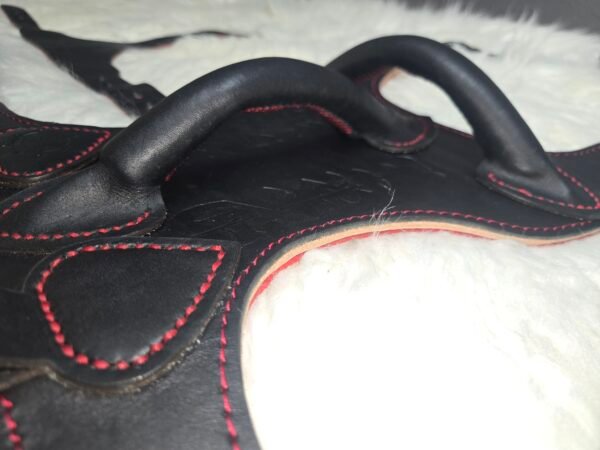 Harness V2.3 Closeup 2 Close-up of the Owned Riding BDSM Harness showing its three-layer leather construction: black 5 oz English Bridle outer, natural 7 oz veg-tan structural core, red 1–2 oz deersplit lining, and raised leather handles for easy grip.