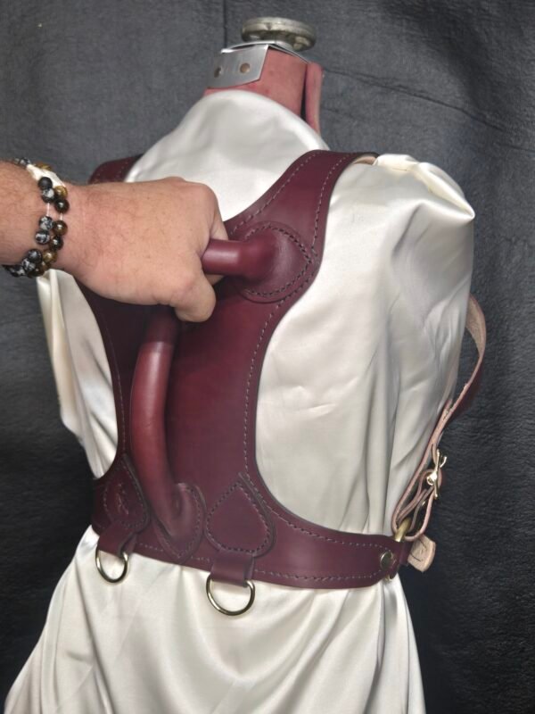 Harness v2.3 - on Mannequin Burgundy - Hand on 1 Burgundy English Bridle leather Owned Riding BDSM Harness with solid brass D-rings, shown from the rear while the horizontal control handle is being gripped.