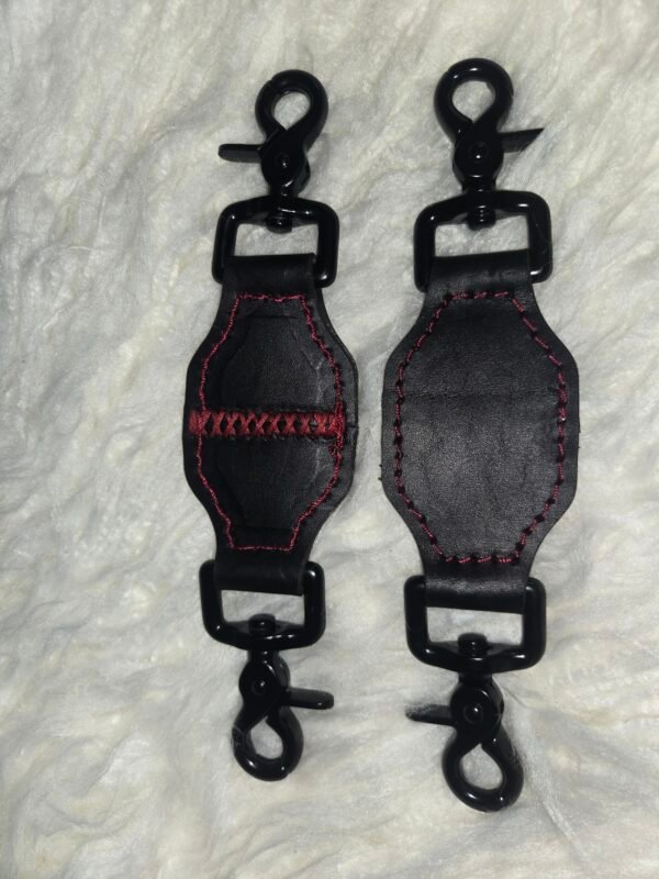Tethers Pair of black English Bridle leather tethers with red stitching and black swivel snap hardware, shown laid flat on a white fur background.